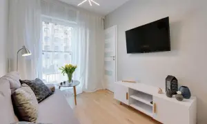 Przytulny salon z sofą, stolikiem i telewizorem. W wazonie żółte tulipany, a na komodzie ozdoby. W tle okno z białymi zasłonami, widok na budynek. Swarożyn.