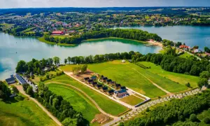 Malowniczy krajobraz z jeziorem i zabudowaniami, w tym domkami letniskowymi. W oddali widać miasteczko, a na pierwszym planie pola i lasy. To idealne miejsce na wypoczynek, jak w bajkowym zamku Lapalice.