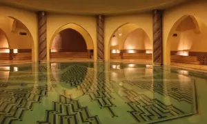 Wnętrze hammamu z basenem o geometrycznym wzorze na dnie. Wokół łuki, kolumny zdobione mozaiką i kamienne ławy.
