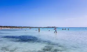 Słoneczny dzień na plaży Es Trenc. Ludzie kąpią się w turkusowej wodzie, niektórzy spacerują po płytkiej wodzie.