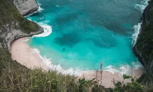 Widok z góry na zatokę Kelingking z turkusową wodą, piaszczystą plażą i stromymi klifami porośniętymi zielenią.