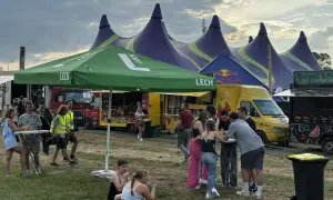 Ludzie bawią się pod namiotami na festynie, przypominającym atmosferę Oktoberfest 2025. W tle food trucki i zielony parasol z logo Lech.