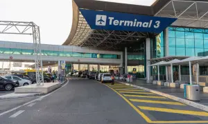 Wejście do Terminalu 3 lotniska Fiumicino. Samochody czekają przed budynkiem, a nad nimi wisi niebieski baner z napisem "Terminal 3".