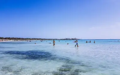 Słoneczny dzień na plaży Es Trenc. Ludzie kąpią się w turkusowej wodzie, niektórzy spacerują po płytkiej wodzie.