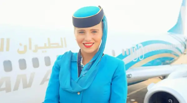 Uśmiechnięta stewardessa w niebieskim mundurku i charakterystycznym nakryciu głowy pozuje przed samolotem.