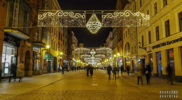 Toruń stare miasto nocą, udekorowane świątecznymi lampkami. Brukowana ulica pełna ludzi, otoczona kamienicami.