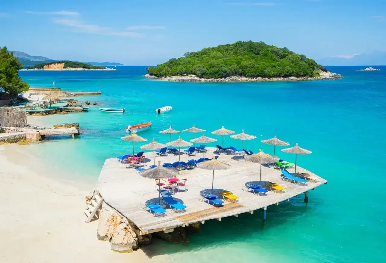 Albania jakie morze? Turkusowa woda, piaszczysta plaża, leżaki i parasole na drewnianym pomoście. Widok na zieloną wyspę.
