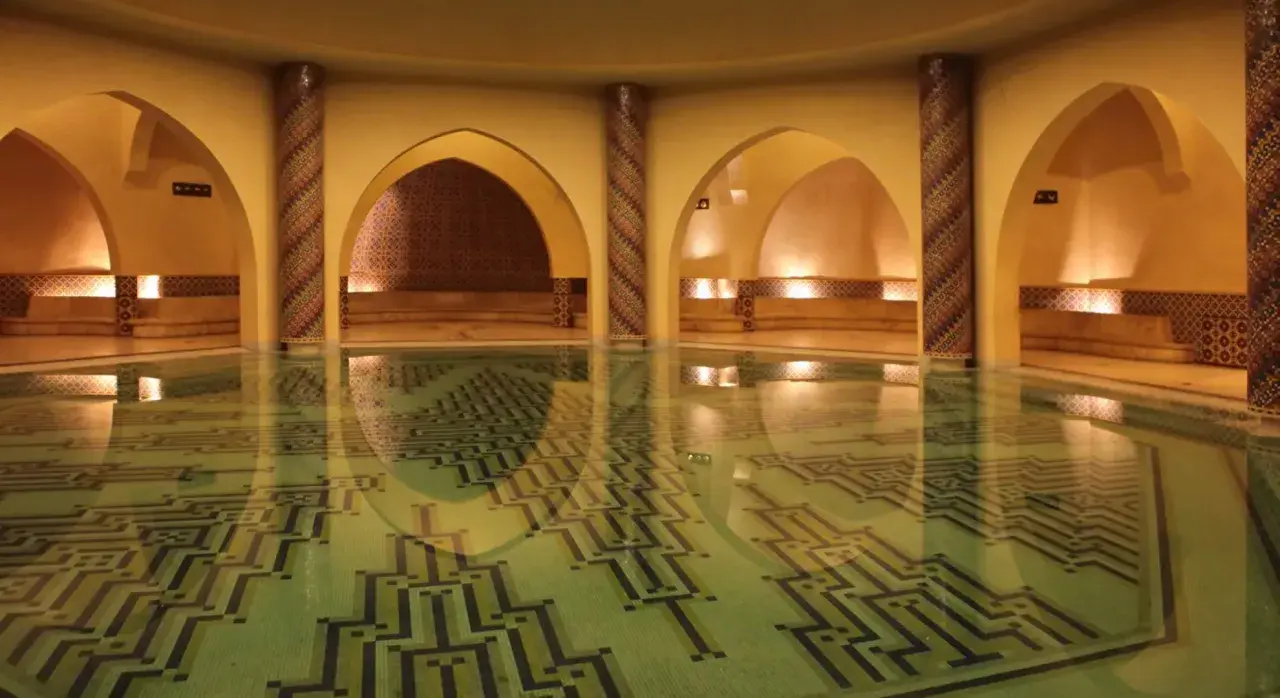Wnętrze hammamu z basenem o geometrycznym wzorze na dnie. Wokół łuki, kolumny zdobione mozaiką i kamienne ławy.