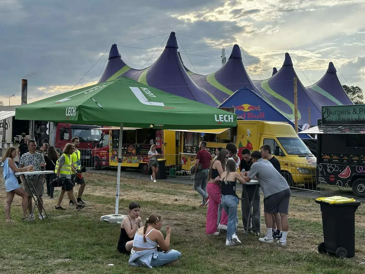 Ludzie bawią się pod namiotami na festynie, przypominającym atmosferę Oktoberfest 2025. W tle food trucki i zielony parasol z logo Lech.