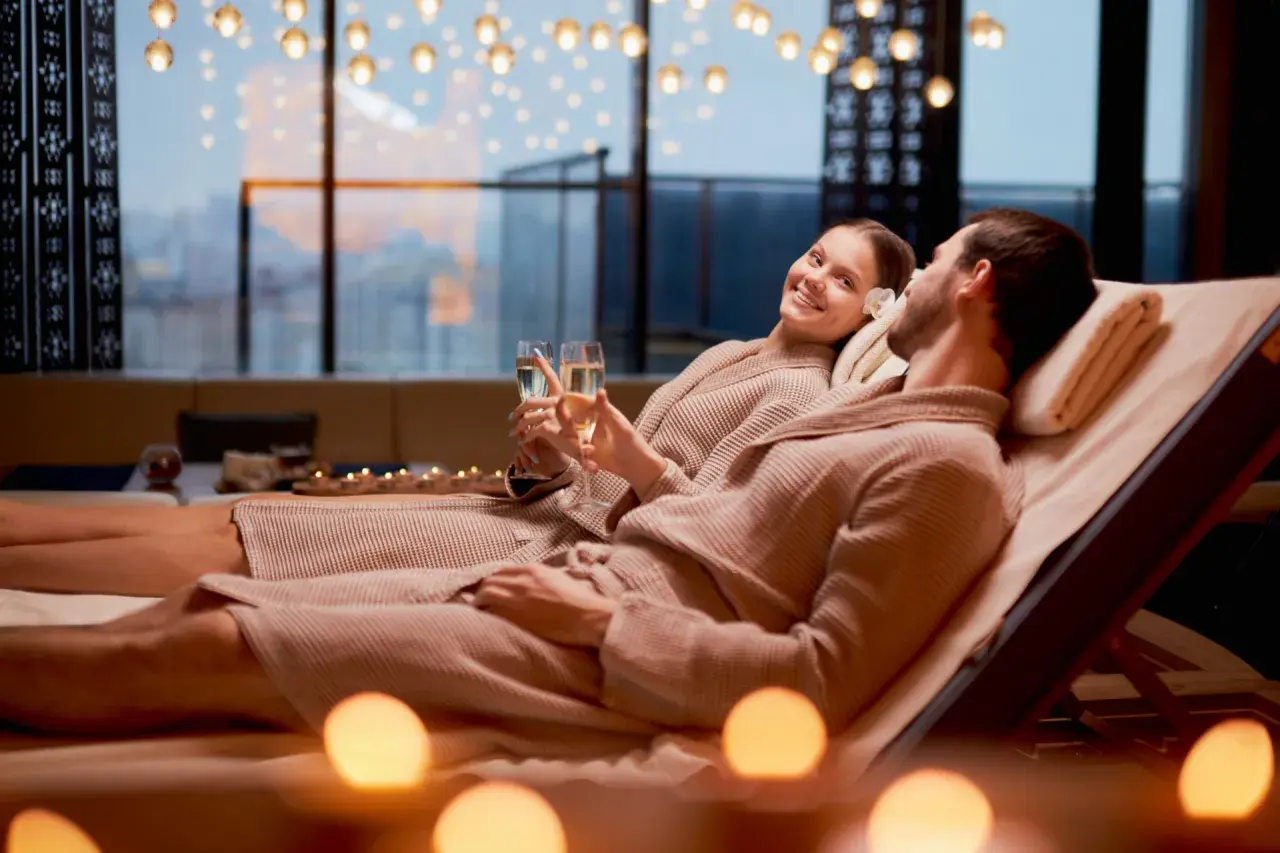 Para relaksu w hotelach dla dorosłych: para w szlafrokach wznosi toasty szampanem, otoczona świecami i widokiem na miasto.