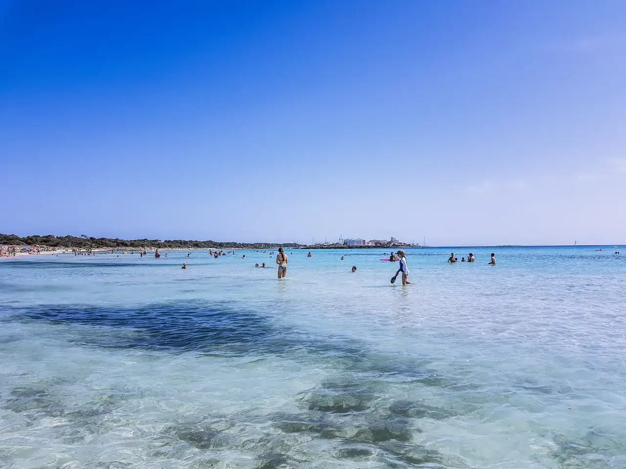 Słoneczny dzień na plaży Es Trenc. Ludzie kąpią się w turkusowej wodzie, niektórzy spacerują po płytkiej wodzie.