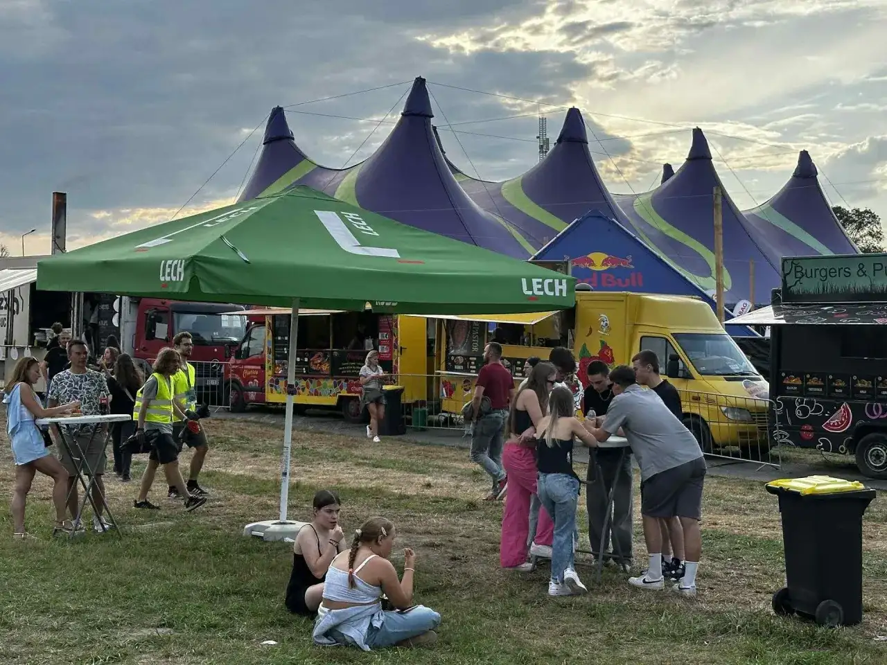 Ludzie bawią się pod namiotami na festynie, przypominającym atmosferę Oktoberfest 2025. W tle food trucki i zielony parasol z logo Lech.