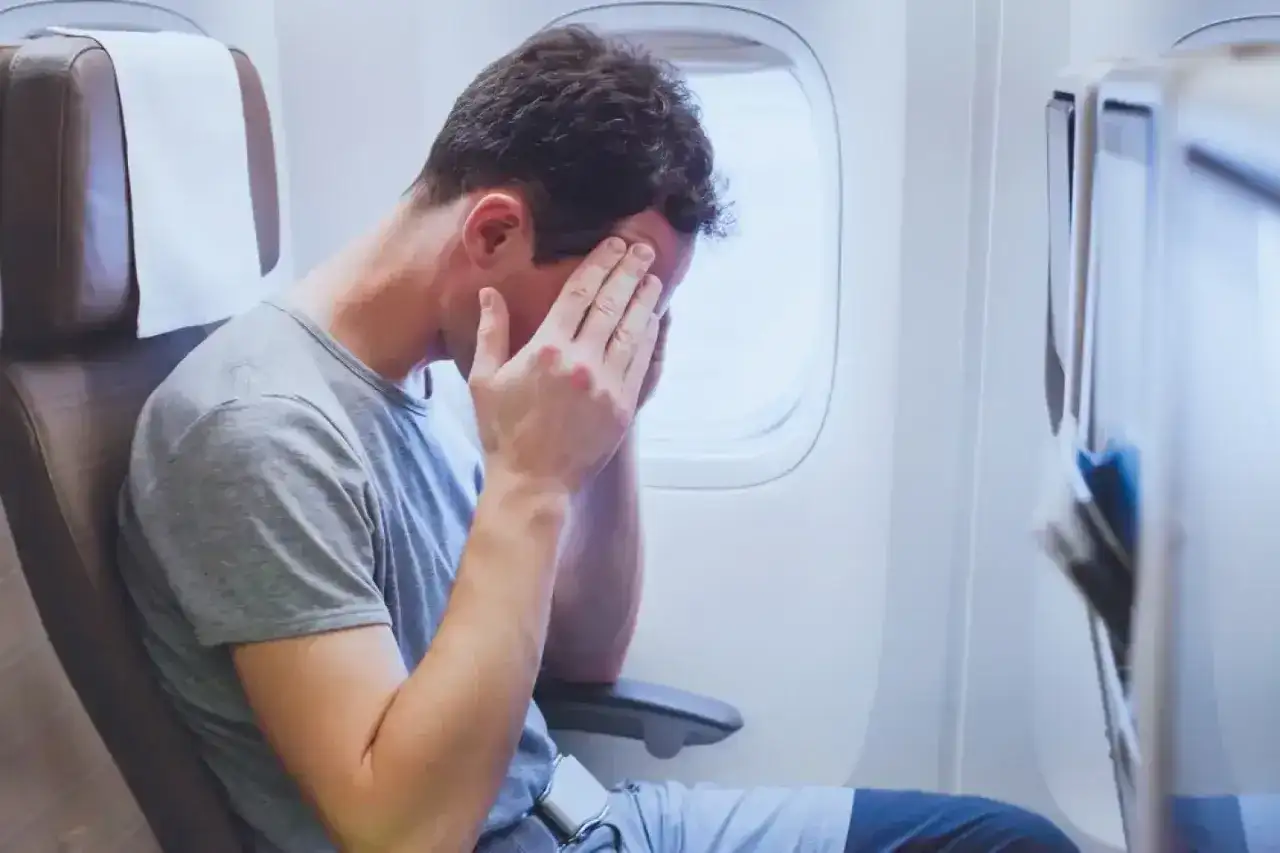 Mężczyzna w samolocie z rękami na twarzy, cierpiący na jetlag. To uczucie zmęczenia i dezorientacji po podróży.