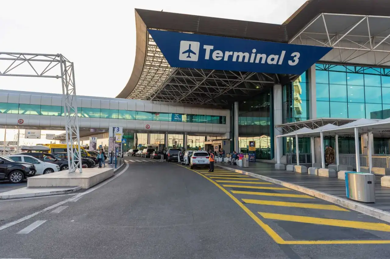 Wejście do Terminalu 3 lotniska Fiumicino. Samochody czekają przed budynkiem, a nad nimi wisi niebieski baner z napisem "Terminal 3".
