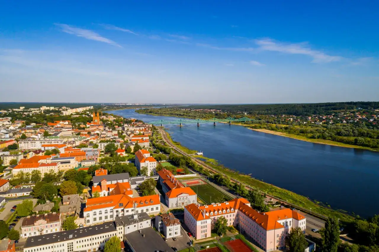 Widok z lotu ptaka na miasto nad szeroką rzeką Wisłą. W oddali zielony most, a bliżej zabudowania z czerwonymi dachami.