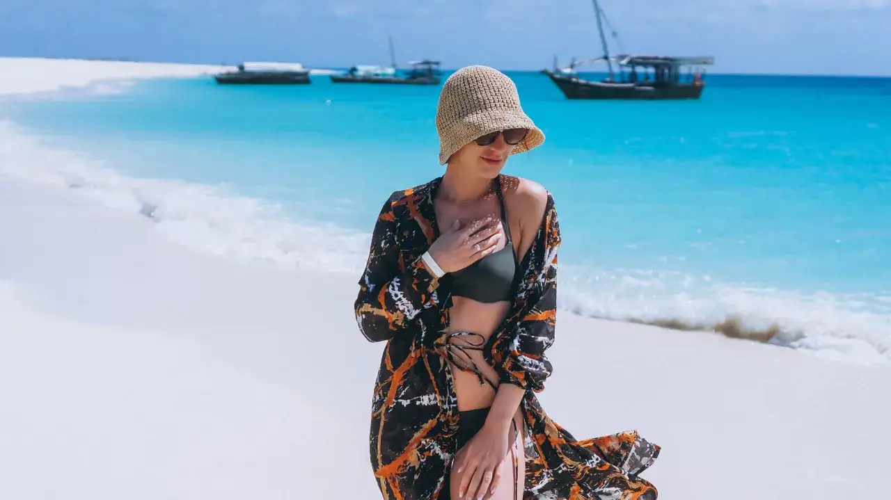 Kobieta w kapeluszu i pareo na plaży, marząca o wakacjach na Zanzibarze. W tle turkusowa woda i łodzie.