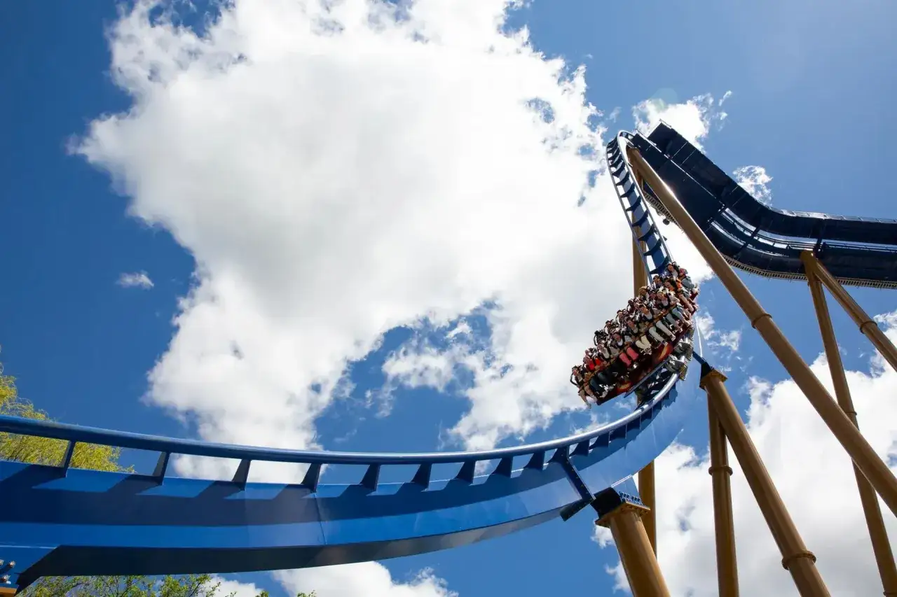 Wagonik rollercoastera z pasażerami pędzi po niebieskich torach na tle błękitnego nieba z chmurami.