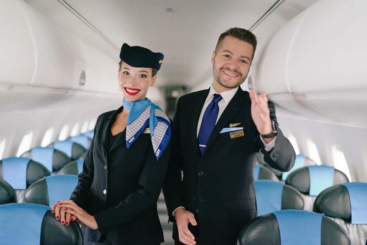 Uśmiechnięta stewardessa i steward w uniformach LOT pozdrawiają pasażerów na pokładzie samolotu.