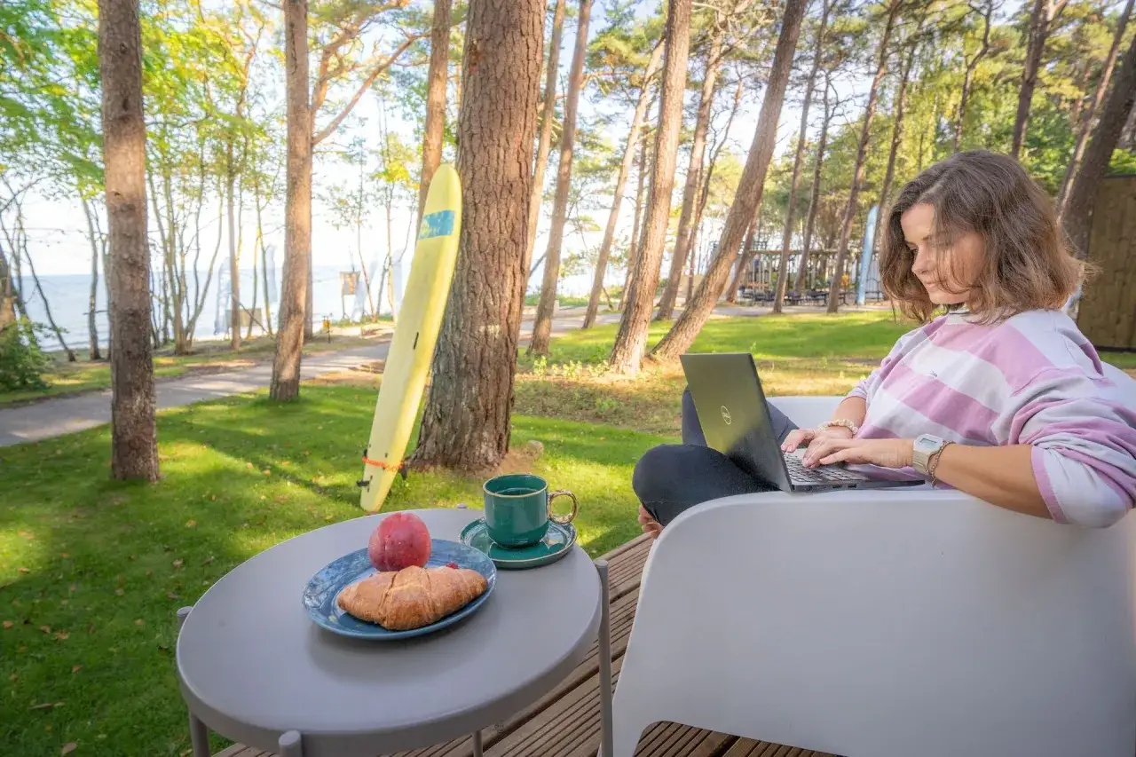 Kobieta pracuje na laptopie przy stoliku z kawą i croissantem. W tle morze i plaża, idealne miejsce na tanie noclegi nad morzem blisko plaży.