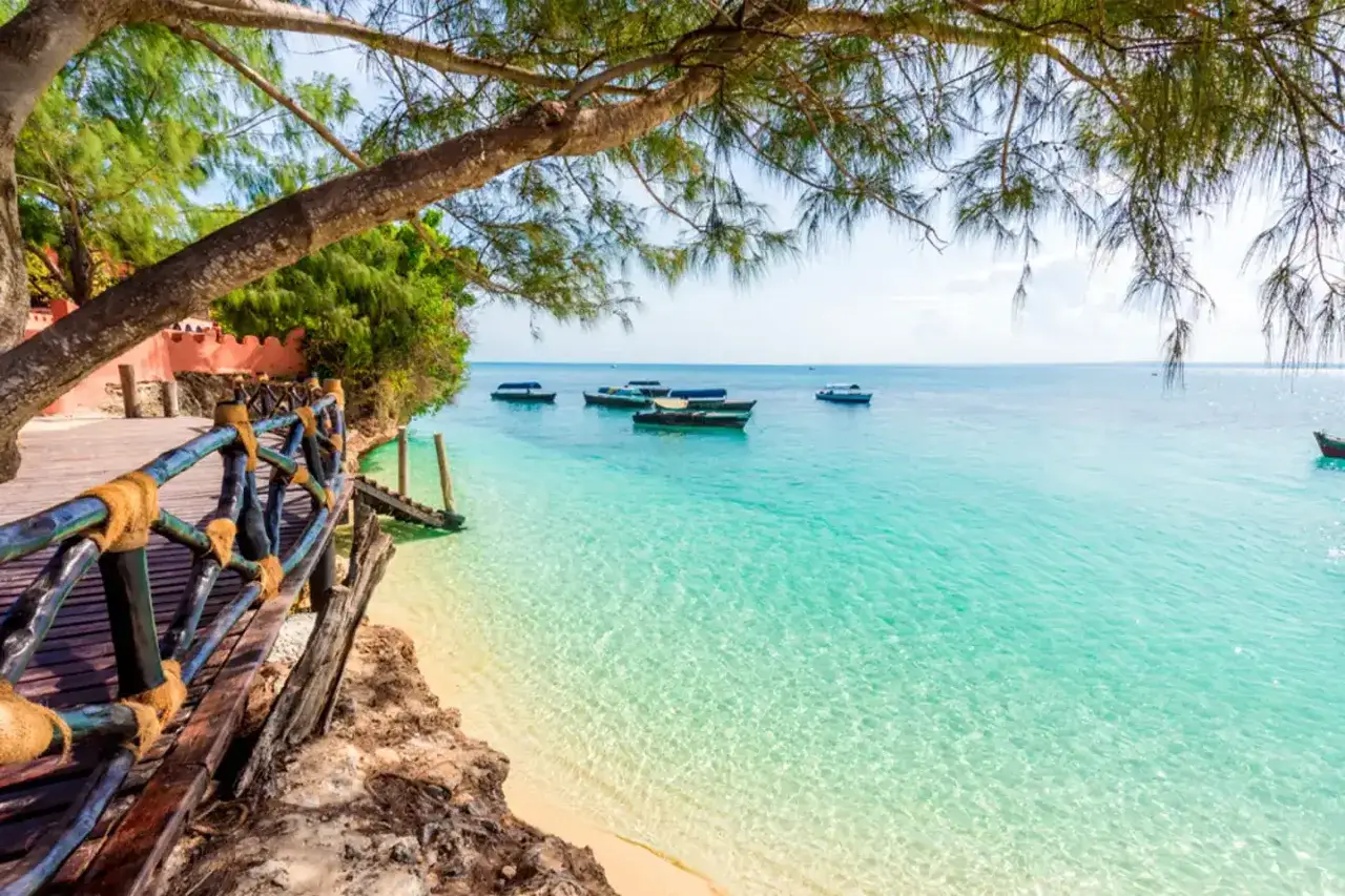 Drewniana kładka prowadzi do turkusowej wody, gdzie kołyszą się łodzie. Idealne miejsce na zanzibar wakacje.