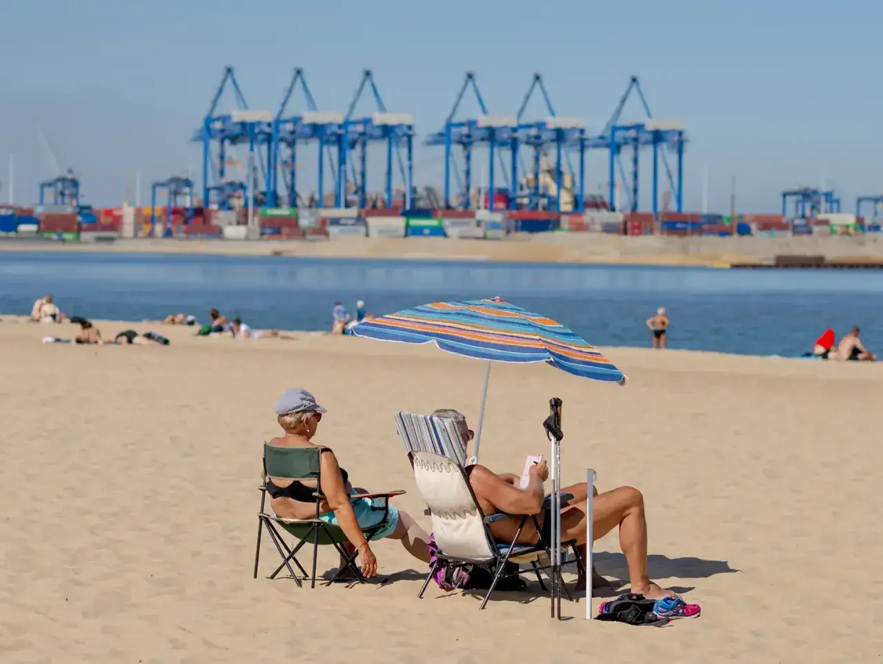 Relaks na jelitkowo plaży. Dwie osoby odpoczywają na krzesłach pod parasolem, w tle portowe dźwigi i kontenery.