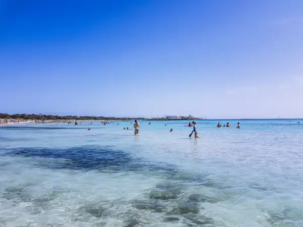 Słoneczny dzień na plaży Es Trenc. Ludzie kąpią się w turkusowej wodzie, niektórzy spacerują po płytkiej wodzie.