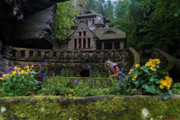 Kamienny domek w skalnym otoczeniu, idealne miejsce na czeska szwajcaria noclegi. Wokół kwitnące kwiaty i zielone drzewa.