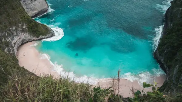 Widok z góry na zatokę Kelingking z turkusową wodą, piaszczystą plażą i stromymi klifami porośniętymi zielenią.